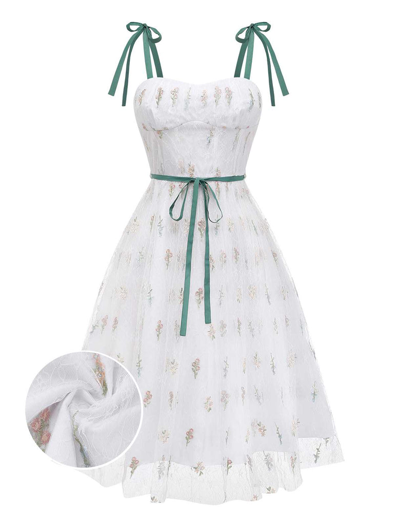 Une délicate robe trapèze en tulle blanc ornée de délicates broderies de fleurs sauvages, de bretelles à nouer en velours vert foncé et d'un ruban de velours assorti à la taille.
